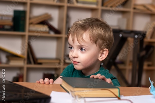 little boy looks into a laptop while sitting at a table with books. close-up portrait of a child. difficulties of distance education. attentive focused look at the monitor.