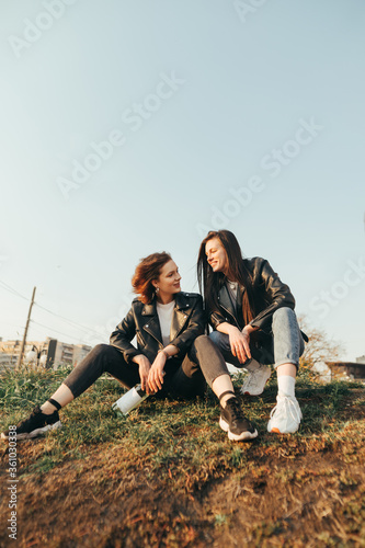 Two happy girls in street clothes are sitting on the grass, holding a bottle of wine, looking at each other and smiling against the background of the sky and cityscape. Girlfriends have fun on a walk