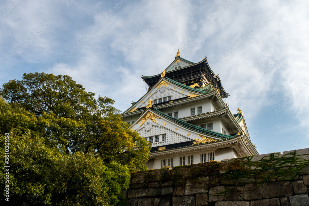 Osaka Castle majestic main tower with green traditional roofs and gold cravings, seen from below with blue sky in the background and trees, autumn, Japan. Osaka Castle majestic main tower with green traditional roofs and gold cravings, seen from below with blue sky in the background and trees, autumn, Japan.