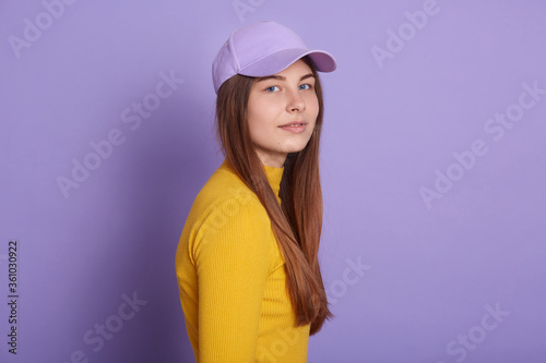 Calm lady dresses casual attire and baseball cap, posing sideways isolated over lilac background, looking directly at camera, has long beautiful hair.