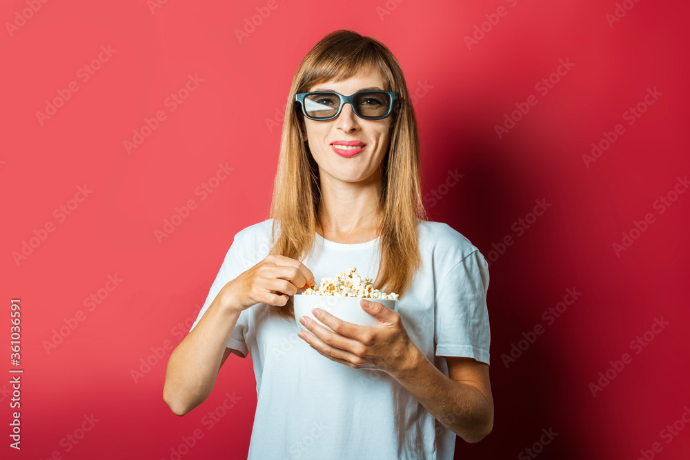 Young beautiful woman in 3D glasses and a bowl of popcorn on a pink background. The concept of watching a movie, movie news, cinema, rating Young beautiful woman in 3D glasses and a bowl of popcorn on a pink background. The concept of watching a movie, movie news, cinema, rating