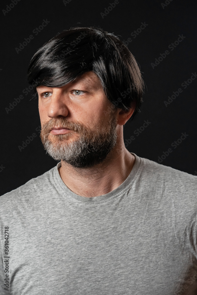 Fototapeta premium Portrait of attractive man in his 40s with grey and black beard and long black hairs. Black background. Male dressed in grey t-shirt. Looking away from the camera.