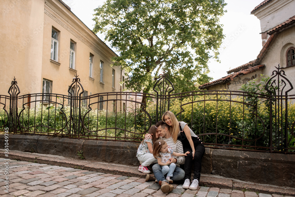 Fototapeta premium Parents in casual clothes with their preschooler daughters hug sitting on the road under a green flowering tree against a yellow field. Mother with blond hair in a jumpsuit hugs her husband.
