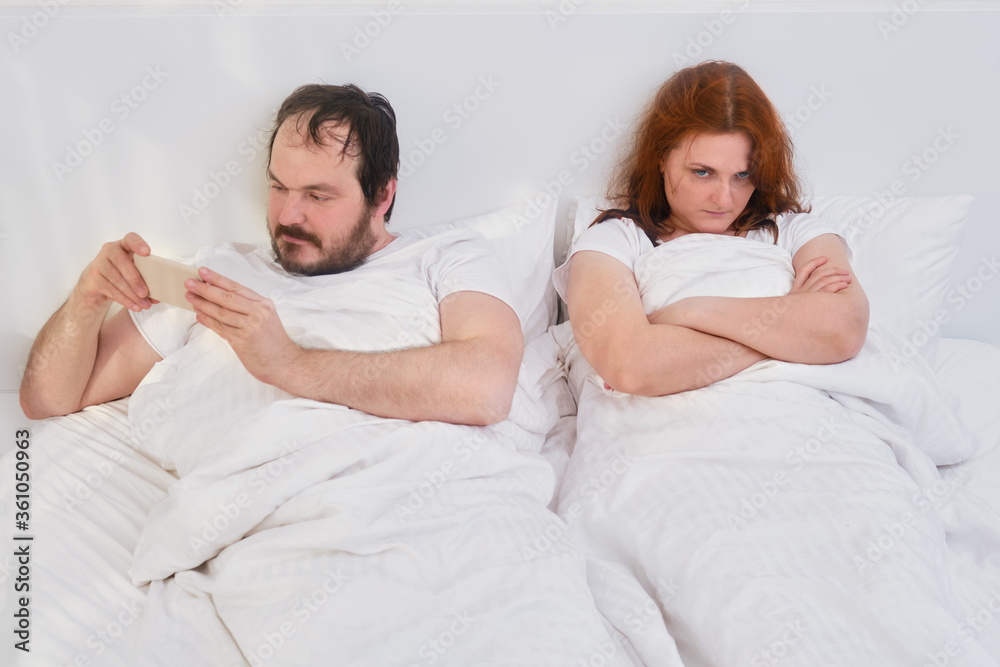 A woman resents a man lying in a white bed. Wife dissatisfied with her husband in the bedroom. Family problems due to quarantine isolation due to the coronavirus pandemic.