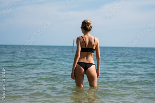 Young woman standing on a beach and enjoying the sun. Young woman in bikini looking at sea water on the beach of tropical resort. Rear view. Vacation and travel concept. © Volodymyr