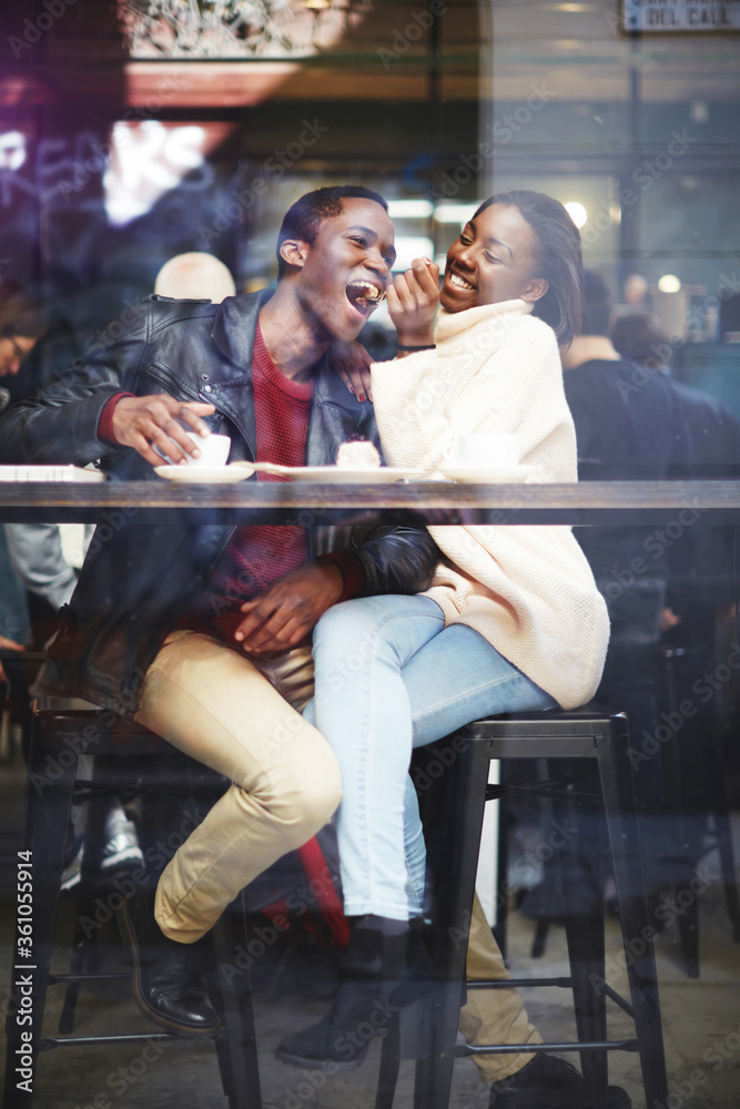 View through cafe window of a young happy dark skinned man and woman having fun while sitting together in bar, cheerful smiling black couple enjoying recreation time while having lunch in coffee shop