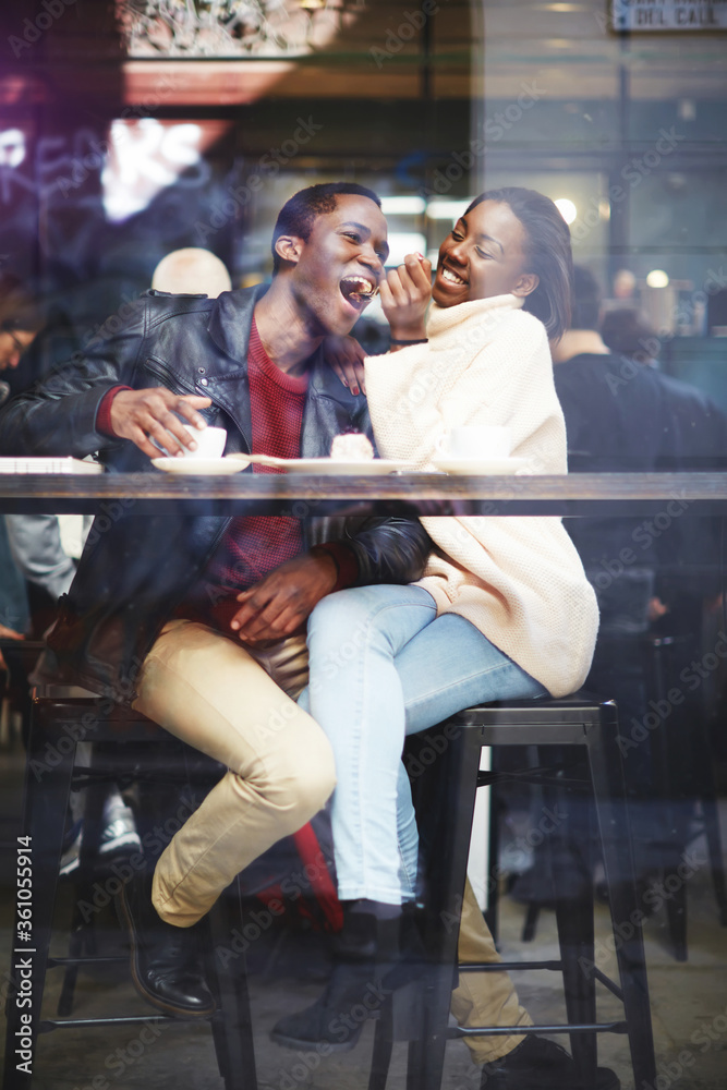 View through cafe window of a young happy dark skinned man and woman having fun while sitting together in bar, cheerful smiling black couple enjoying recreation time while having lunch in coffee shop View through cafe window of a young happy dark skinned man and woman having fun while sitting together in bar, cheerful smiling black couple enjoying recreation time while having lunch in coffee shop