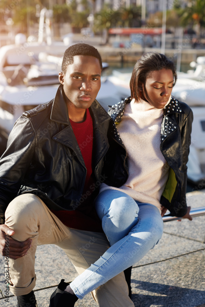 Portrait young fashionable professional models posing outdoors, stylish black couple sitting together in beautiful yacht port of Barcelona