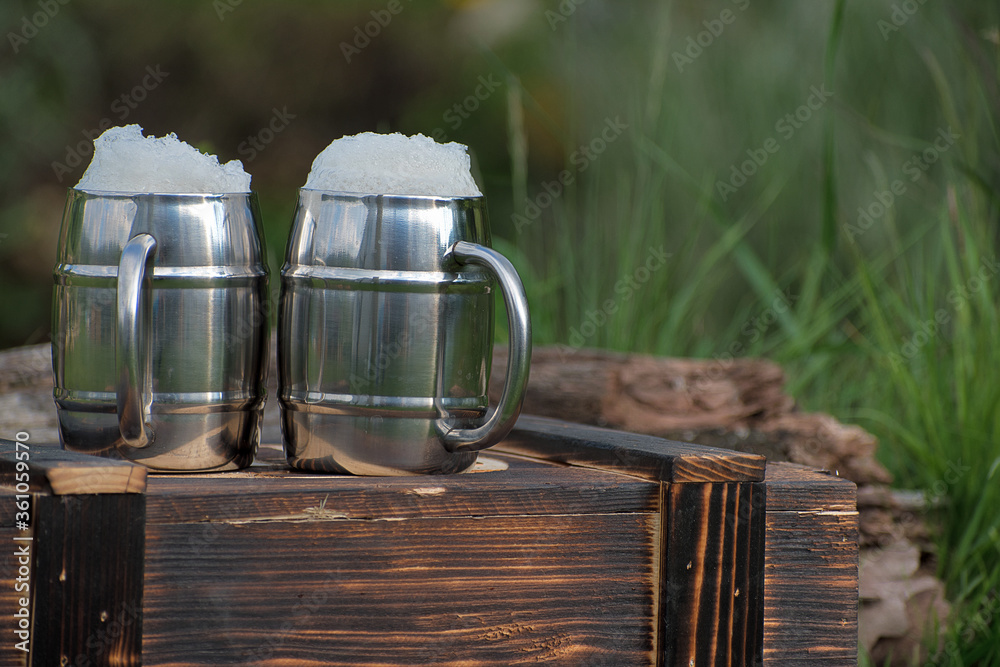 Wooden charred box. Two mugs with beer, white beer foam. Flame tanned wood. Blurred garden background. Wooden charred box. Two mugs with beer, white beer foam. Flame tanned wood. Blurred garden background.