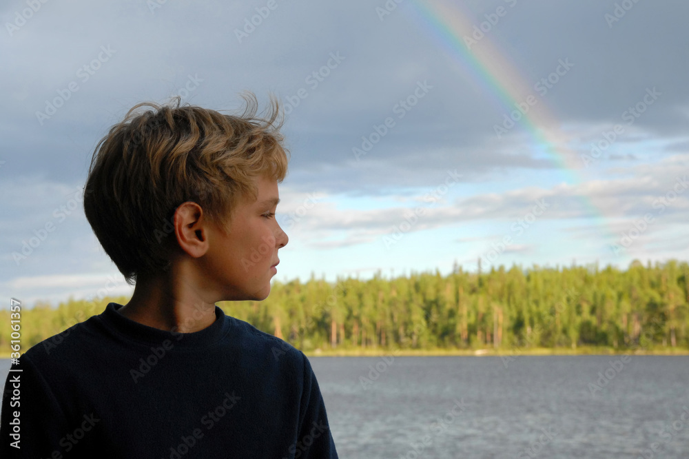 Fototapeta premium Cute blonde boy on the lake with rainbow above it. Outdoor travel. Republic of Karelia, Russia.