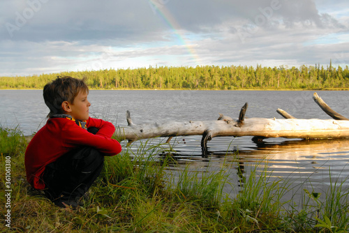Cute boy on the lakeshore with rainbow above it. Outdoor travel. Republic of Karelia, Russia. photo