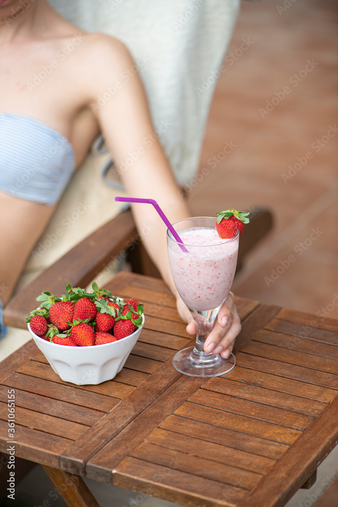 Woman on a sunbed holding a strawberry smoothie next to a bowl of strawberries