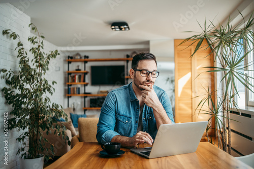 Portrait of a casual businessman thinking while looking at laptop screen, keeping hand on chin. photo