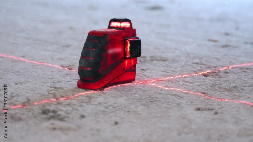 Close-up of modern engineering device set on the floor, installation of laser level for measurement on renovation site. Laser red rays creating horizontal crosshair for accurate measures on Close-up of modern engineering device set on the floor, installation of laser level for measurement on renovation site. Laser red rays creating horizontal crosshair for accurate measures on