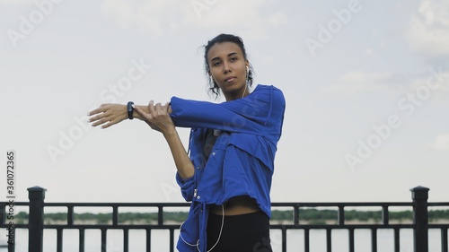 Young Afro-american woman in headphones performs stretching and warming before running in the park