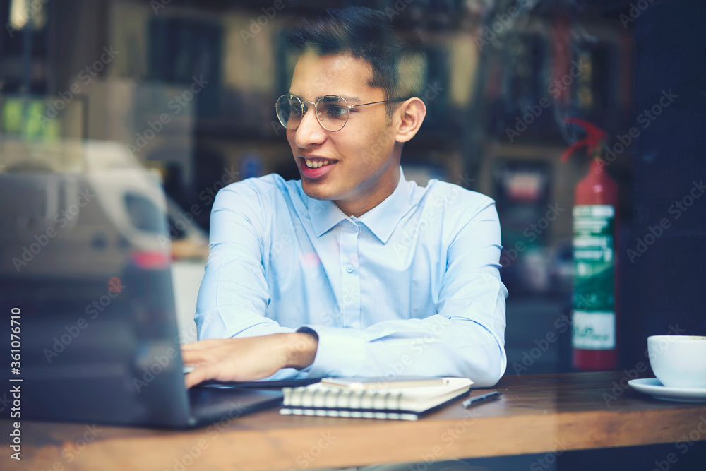 Fototapeta premium Positive young male student in stylish eyeglasses watching funny video on laptop computer connected to internet.Smiling man reading news in websites and browsing information sitting in cafeteria.