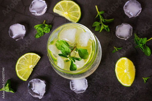 Lime mojito in transparent glass on dark background. Around - pieces of fresh lime, ice and mint. Top view, flat lay, minimalism. The concept of summer citrus refreshing drinks, cocktails, lemonade
