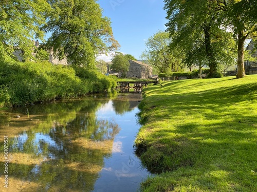 Linton Beck, with a green grass sloping bank, old trees and a footbridge, over reflective water in, Linton, Skipton UK