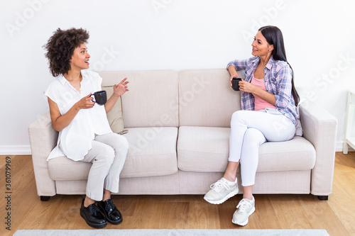 Two female friends in social distancing sitting on sofa in coronavirus pandemic time. Best friends having coffee together while separated by social distancing on sofa at home