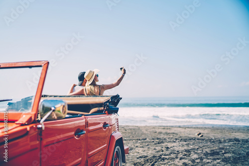 Romantica couple making photo on smartphone camera standing near vintage rental car on ocean beach enjoying summer vacation together  hipsters taking picture on cellular resting near sea on weekends