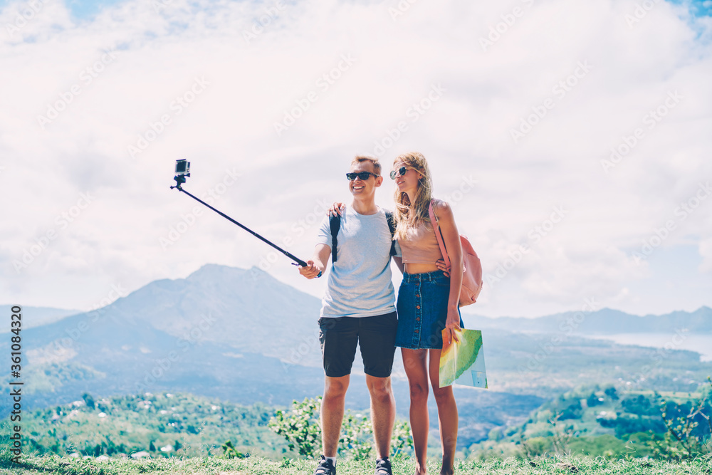 Hipster couple using monopod and smartphone camera taking picture during journey in rural environment,male and female tourist making photo standing on highland in tropical environment resting together