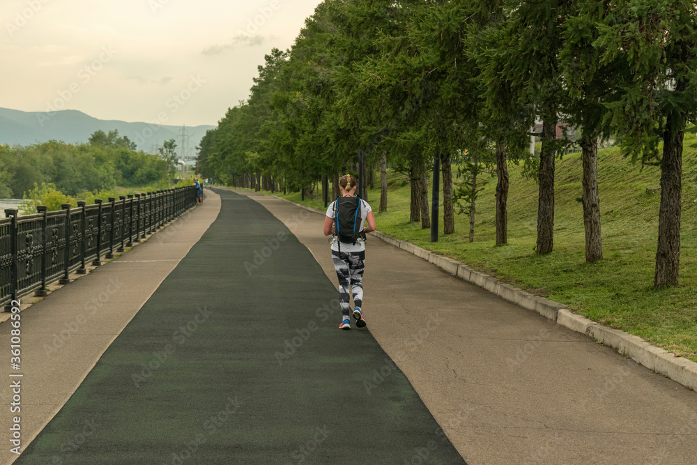 Girl runs along promenade. View from the back. Backpack, sportswear. Alley of green larches. Concept of a healthy lifestyle, tourism, hiking.