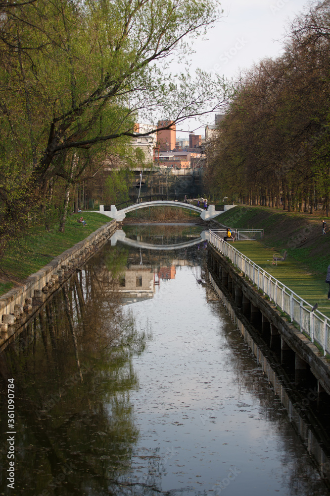 MOSCOW / RUSSIA - 20/04/2019 narrow water Canal in the Krasnaya Presnya park with people relaxing at the quay, a bridge and buildings, tall trees with branches, green leaves in the background