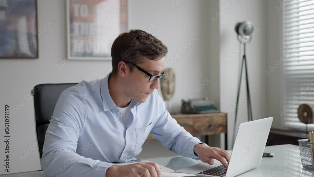 young brunet man in glasses and blue shirt sits at table with laptop in office. is attentive, serious and busy. works multitasking, hurries. types on laptop, makes notes, uses calculator. young brunet man in glasses and blue shirt sits at table with laptop in office. is attentive, serious and busy. works multitasking, hurries. types on laptop, makes notes, uses calculator.
