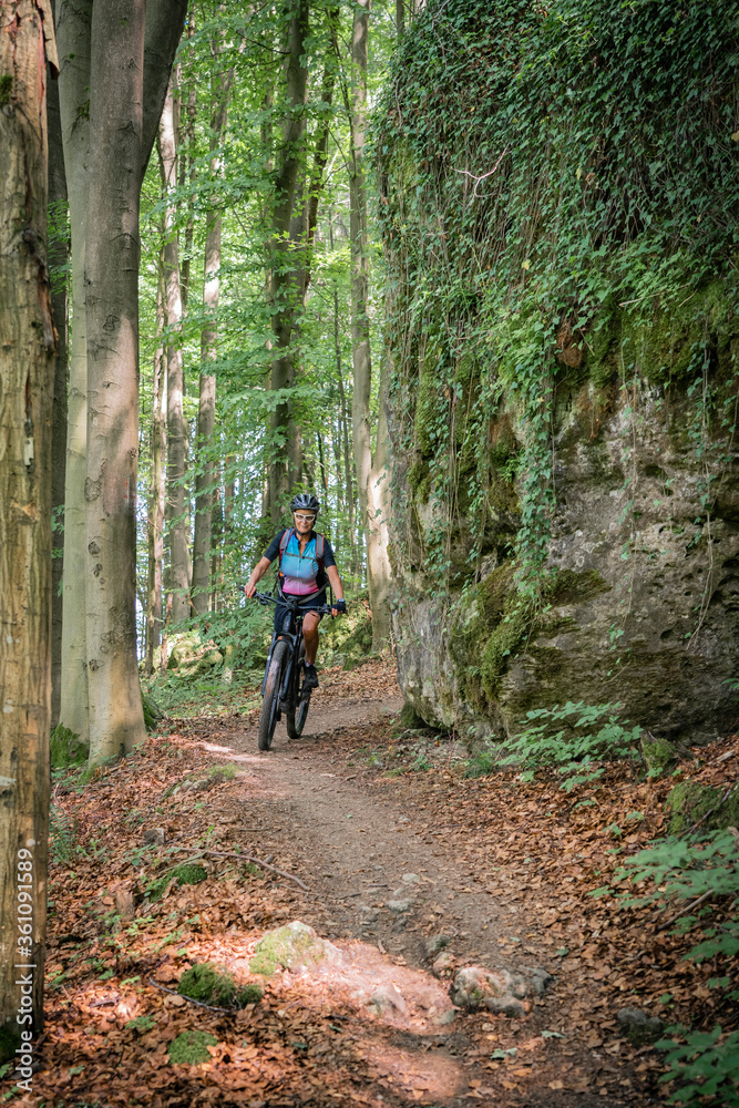 pretty senior woman underway on her electric mountain bike on a rocky forest trail in Franconian Switzerland, Bavaria, Germany