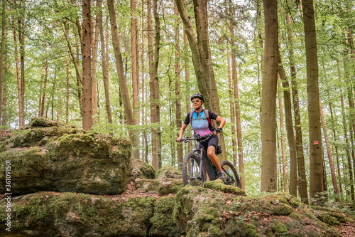 pretty senior woman underway on her electric mountain bike on a rocky forest trail in Franconian Switzerland, Bavaria, Germany