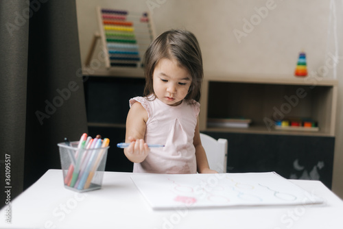 Playful beautiful creative little girl is painting with felt-tip pens on paper at table in children's room.