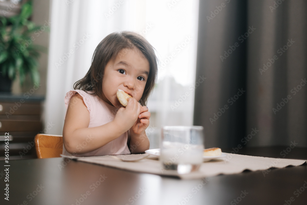 Pretty beautiful little two-year-old girl wearing white dress is eating cheesecake for breakfast at the table in kitchen room. Glass of milk on the table. Pretty beautiful little two-year-old girl wearing white dress is eating cheesecake for breakfast at the table in kitchen room. Glass of milk on the table.