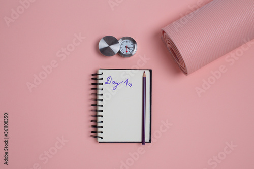 Top view of yoga mat with alarm clock, pencil and Day 1 written on notebook page. Pink background with copy space