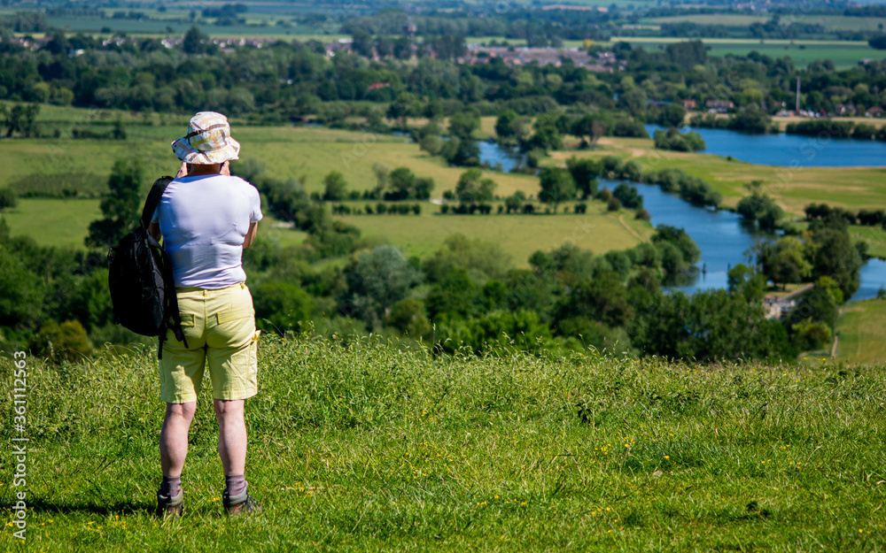 An older woman photographing a landscape in Wittenham Clumps, carrying backpack, enjoying the weather, lady wearing blue t-shirt, creamy shorts and hat, countryside stroll with river view