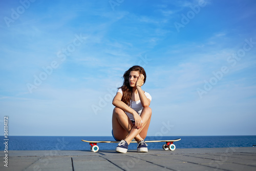 Beautiful sexy girl sits on longboard with amazing horizon of sea on background, young hipster girl posing at beautiful sunset light sitting on her skateboard, attractive woman enjoying sunny evening photo