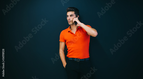 Serious caucasian trendy dressed hipster guy standing near black wall promotional background talking on mobile phone,confident businessman making cellular call posind in stylish outfit indoors