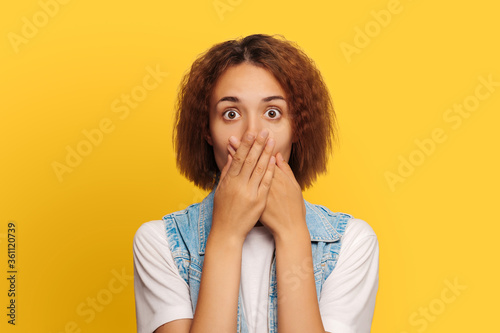 Portrait of a surprised, shocked girl with curly hair, covers her mouth with her hands, look at the camera, dressed in a white T-shirt and jeans, isolated on a yellow background