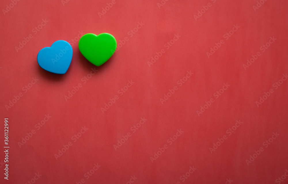 Fototapeta premium Two blue and green hearts lying close together isolated on right corner of red background. Flat lay with empty space for text.