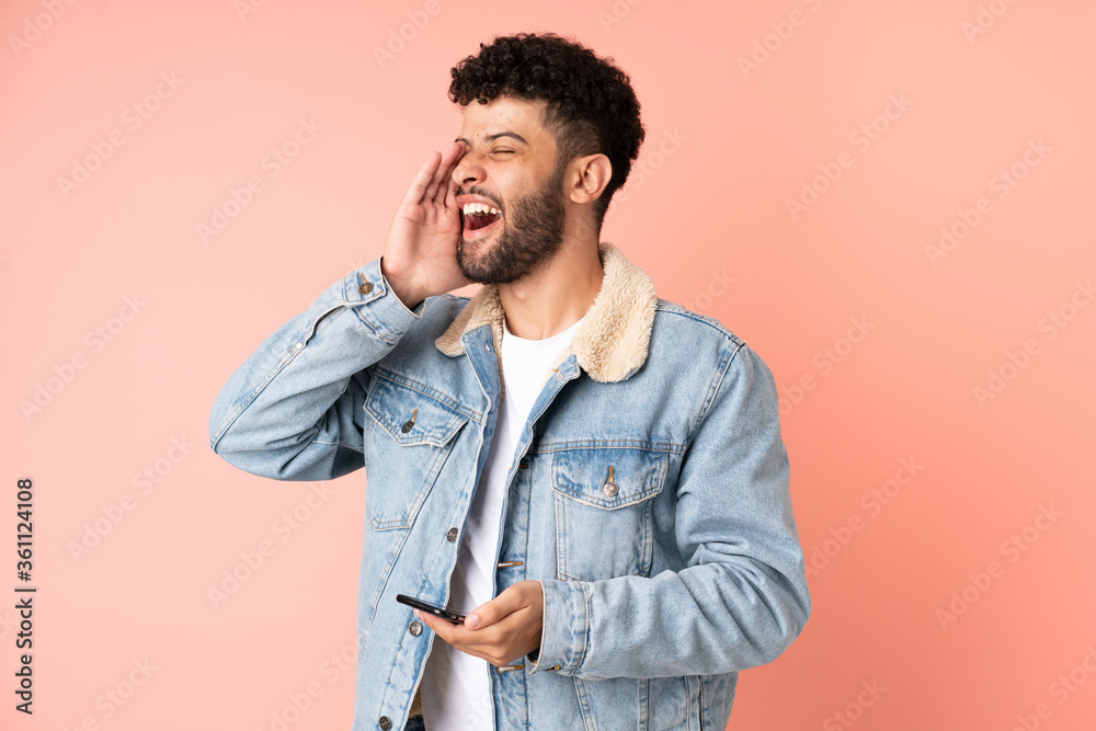 Young Moroccan man using mobile phone isolated on pink background shouting with mouth wide open to the side Young Moroccan man using mobile phone isolated on pink background shouting with mouth wide open to the side