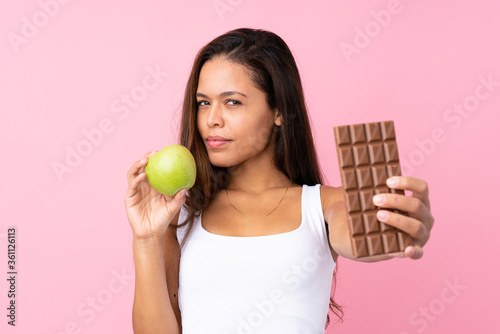 Young Brazilian girl over isolated pink background taking a chocolate tablet in one hand and an apple in the other