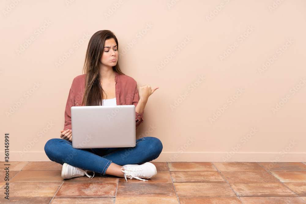 Teenager student girl sitting on the floor with a laptop unhappy and pointing to the side Teenager student girl sitting on the floor with a laptop unhappy and pointing to the side