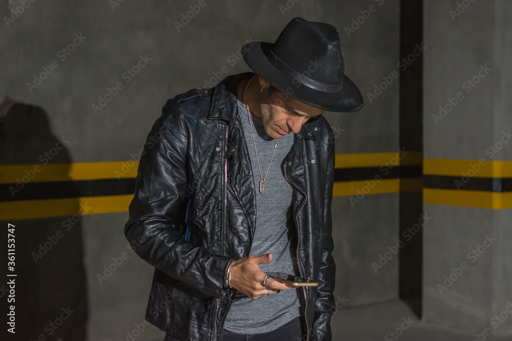 Latin man with black leather jacket and hat looking his smartphone in a underground car parking Latin man with black leather jacket and hat looking his smartphone in a underground car parking