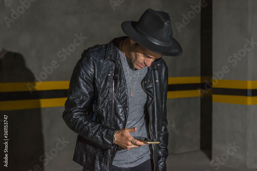 Latin man with black leather jacket and hat looking his smartphone in a underground car parking