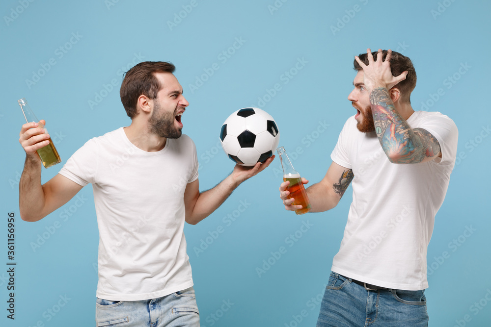 Irritated nevous young men guys friends in white t-shirt isolated on pastel blue background. Sport leisure concept. Cheer up support favorite team with soccer ball, beer bottle looking at each other.