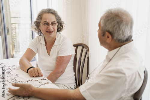 Old aged, Turkish, happy senior couple enjoying time together, looking to each other, laughing, sitting in living room on chair. 