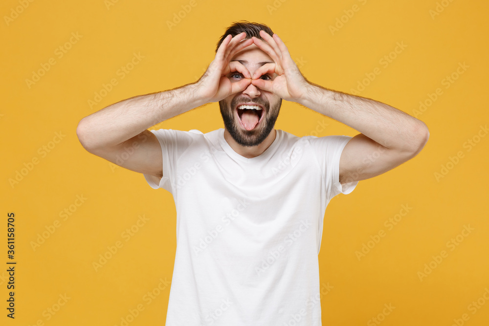 Funny young bearded guy in white casual t-shirt posing isolated on yellow background. People lifestyle concept. Mock up copy space. Hold hands near eyes imitating glasses or binoculars showing tongue. Funny young bearded guy in white casual t-shirt posing isolated on yellow background. People lifestyle concept. Mock up copy space. Hold hands near eyes imitating glasses or binoculars showing tongue.