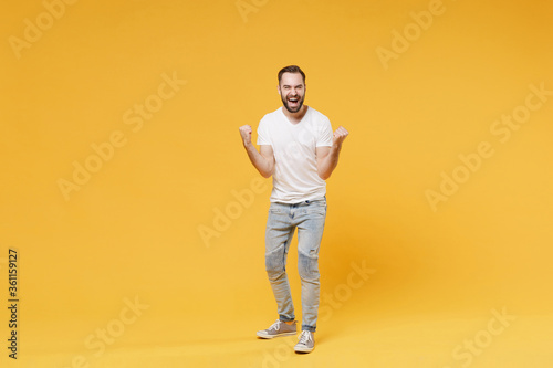 Joyful young bearded man guy in white casual t-shirt posing isolated on yellow wall background studio portrait. People sincere emotions lifestyle concept. Mock up copy space. Doing winner gesture.