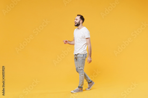 Side view fo cheerful young bearded man guy in white casual t-shirt posing isolated on yellow background studio portrait. People emotions lifestyle concept. Mock up copy space. Walking, looking aside.