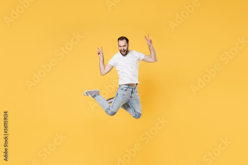 Funny young bearded man guy in white casual t-shirt posing isolated on yellow background studio portrait. People sincere emotions lifestyle concept. Mock up copy space. Jumping showing victory sign.
