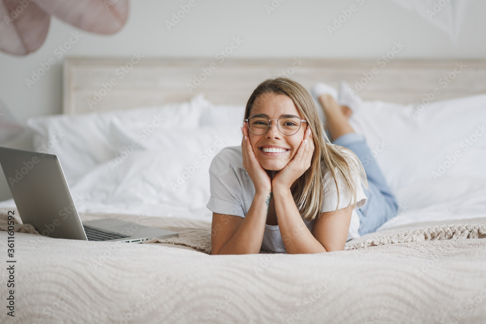 Smiling young woman girl in white t-shirt glasses lying in bed with white sheet pillow blanket spending time in bedroom at home. Rest relax good mood lifestyle concept. Put hands prop up on cheeks. Smiling young woman girl in white t-shirt glasses lying in bed with white sheet pillow blanket spending time in bedroom at home. Rest relax good mood lifestyle concept. Put hands prop up on cheeks.